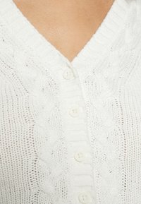 Weißer gestrickter Cardigan mit V-Ausschnitt, Zopfmuster und fünf weißen Knöpfen auf der Vorderseite. Die Textur wirkt weich und atmungsaktiv.