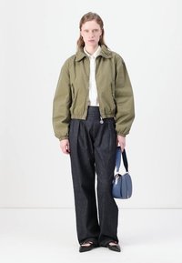 Giacca bomber verde oliva corta, camicia bianca con colletto, pantaloni in denim scuri a vita alta, ballerine nere e borsa blu.
