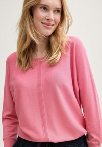 Pulover tricotat cu mânecă lungă, de culoare roz, cu decolteu rotund, având accente rigate pe centru și o croială lejeră. Combinat cu pantaloni negri.