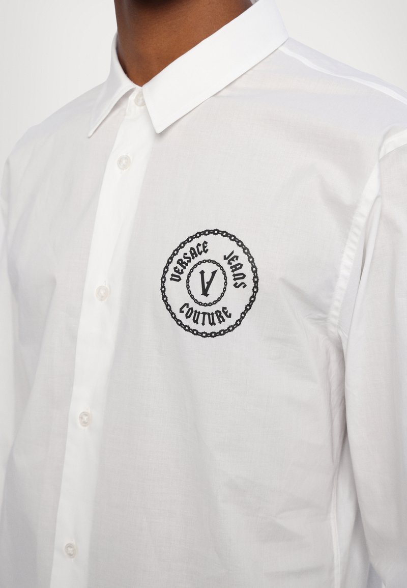 Biała bawełniana koszula z czarnym haftowanym logo w okrągłym wzorze na lewej piersi. Klasyczny kołnierzyk, zapięcie na guziki z przodu oraz fakturowany materiał.