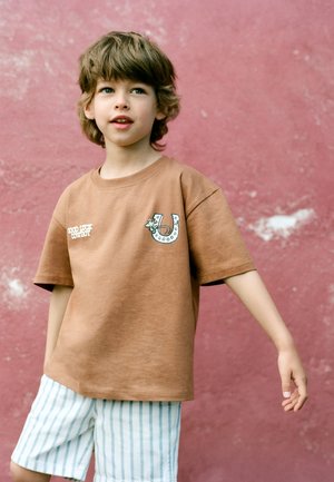 Jong kind met lichtbruin haar dat een bruin "Good Luck Cowboy" shirt en witte gestreepte korte broek draagt, staat tegen een gestructureerde roze muur.