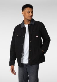 Svart denimjakke med kontrasterende grå krage, trykknapper og frontlommer. Brukes over en hvit skjorte, kombinert med mørke jeans.