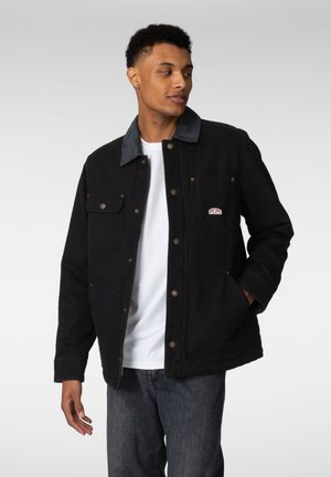 Svart denimjakke med kontrasterende grå krage, trykknapper og frontlommer. Brukes over en hvit skjorte, kombinert med mørke jeans.