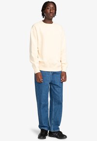 Crèmewit sweatshirt met geribbelde manchetten en zoom, gecombineerd met losvallende blauwe spijkerbroek en zwarte sneakers. Eenvoudig en casual ontwerp.