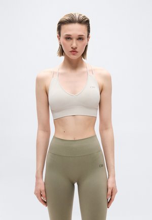 Nainen, jolla on beige urheilurintaliivit ja korkeavyötäröiset oliivinvihreät legginsit, seisoo rentoine käsivarsineen yksiväristä vaaleaa taustaa vasten.