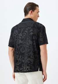 Camisa negra de manga corta con un patrón en relieve de espirales. Tela ligera y semitransparente con un ajuste relajado, que presenta un cuello clásico y costuras traseras.