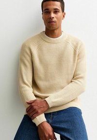 Beige Rippstrickpullover mit Rundhalsausschnitt und Raglanärmeln, kombiniert mit blauen Jeans. Die Textur ist deutlich erhaben und weich.