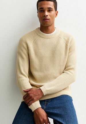 Maglione a coste beige con collo rotondo e maniche raglan. Indossato con jeans in denim blu, esposto su uno sfondo neutro.