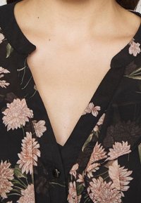 Svart blommig blus med ljusrosa och gröna blommönster, med V-hals och små svarta knappar. Mjuk, lättvikts tyg.