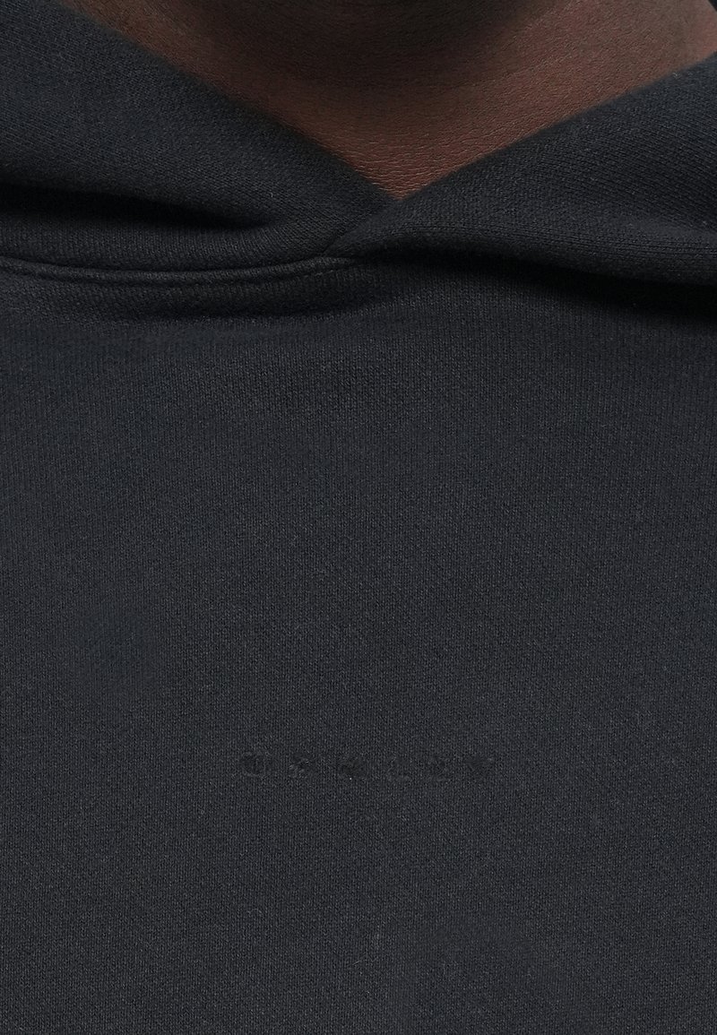 Felpa nera con tessuto texturizzato, dotata di tasca a marsupio anteriore e branding discreto vicino al colletto. Dettagli con cappuccio regolabile visibili.