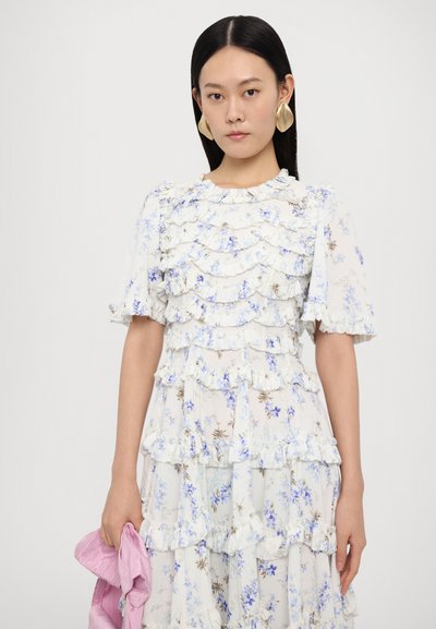 Bloemenjurk in wit met blauwe en bruine patronen, met ruchelagen en korte pofmouwen. Dun, lichtroze accessoire in de hand.