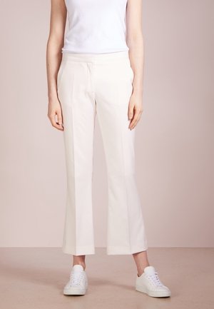 Vrouw draagt witte cropped broek met rechte pijpen, witte mouwloze top en witte sneakers, staand tegen een effen neutrale achtergrond.