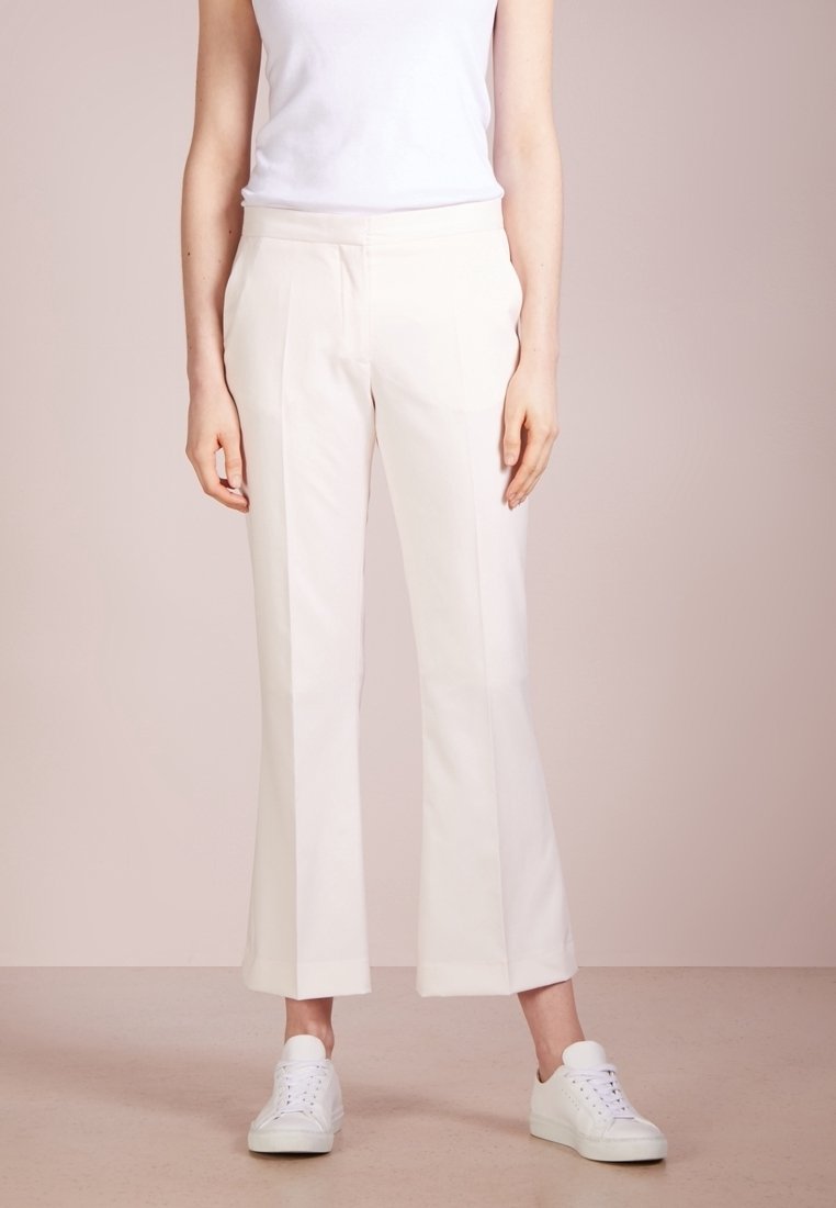 Femme portant un pantalon blanc raccourci droit, un top blanc sans manches et des baskets blanches, debout devant un fond neutre uni.