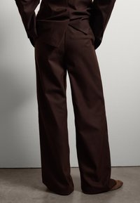 Pantalones marrones a medida hechos de una tela suave, con un diseño de pierna ancha y un detalle de abertura en la parte trasera, combinados con zapatos marrones.