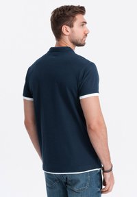 Námornícky polo tričko s bielými akcentmi na rukávoch, žebrovaným golierom, krátkymi rukávmi a těsným střihem, nošené s modrými džíny.