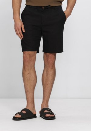 Mand iført sorte shorts med snøre og sorte sandaler, stående med den ene hånd i lommen mod en ensfarvet grå baggrund.