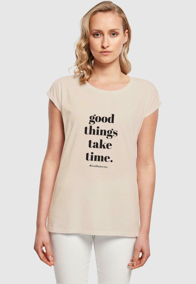 T-shirt beige chiaro con maniche a cappuccio arrotolate, con un testo nero audace che recita "le cose belle richiedono tempo" centrato sulla parte anteriore.