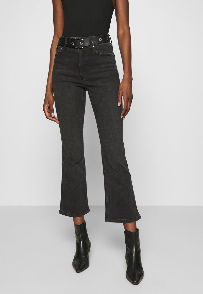 Femme portant un jean flare court noir avec une ceinture noire, un top noir sans manches et des bottines noires pointues, debout devant un fond uni.
