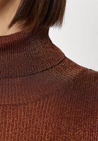 Gros plan d'un pull à col roulé côtelé marron avec une subtile brillance métallique porté par une personne aux cheveux bruns raides.