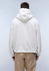 Felpa bianca in pile con superficie testurizzata, cappuccio con coulisse e vestibilità rilassata, abbinata a pantaloni beige chiaro.