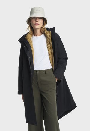 Femme portant un chapeau bob gris clair, un t-shirt blanc, un manteau noir avec doublure beige et un pantalon vert olive, debout devant un fond blanc.