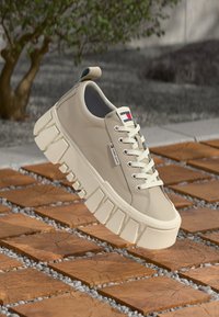 Beige Sneaker mit einer dicken, strukturierten Sohle, glattem synthetischen Obermaterial und Schnürdesign. Verfügt über ein Logo-Tag an der Seite.