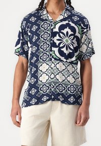 Camicetta a maniche corte con base blu navy, caratterizzata da modelli floreali e geometrici in bianco e verde. Tessuto leggero, vestibilità rilassata.