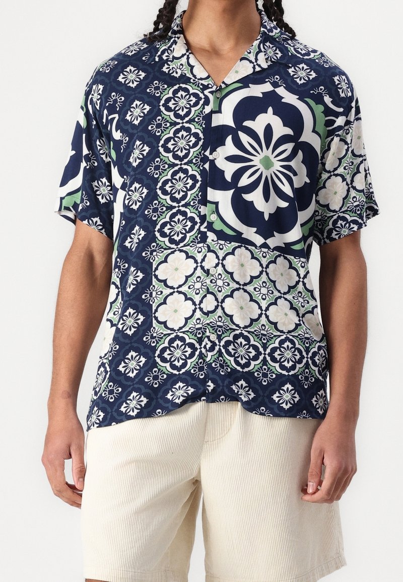 Camicetta a maniche corte con base blu navy, caratterizzata da modelli floreali e geometrici in bianco e verde. Tessuto leggero, vestibilità rilassata.