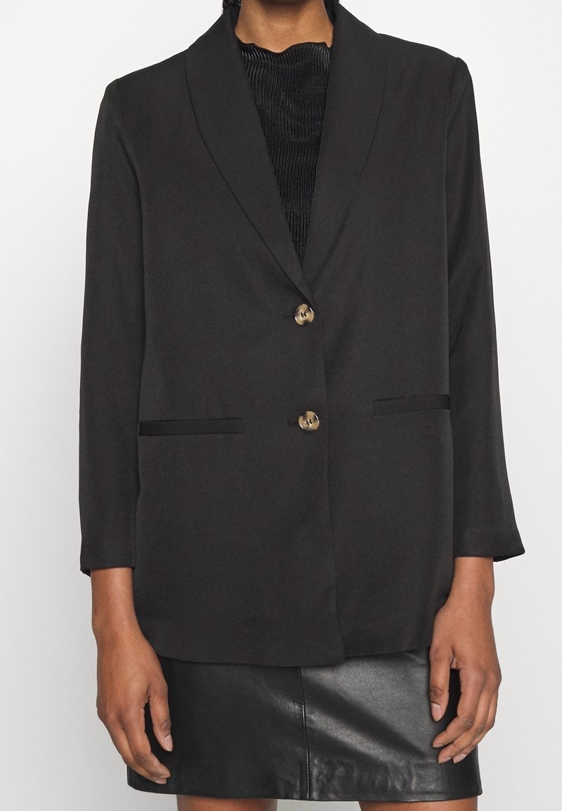 Femme portant un blazer noir à deux boutons sur un top noir texturé et une jupe en cuir noire sur fond blanc uni.