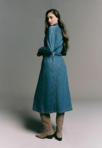 Denimkjole med lange ærmer, bælte i taljen og ankellang nederdel. Har klassisk syning og en lys vasket tekstur. Støvlerne er i beige ruskind.