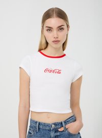 T-shirt bianco cropped con colletto rosso, che presenta il logo "Enjoy Coca-Cola" in carattere corsivo rosso. Abbinato a pantaloni corti di jeans a vita alta.