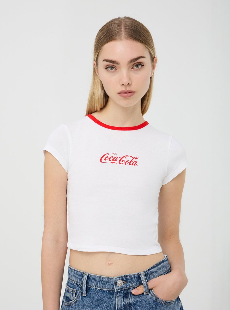 T-shirt bianco cropped con colletto rosso, che presenta il logo "Enjoy Coca-Cola" in carattere corsivo rosso. Abbinato a pantaloni corti di jeans a vita alta.