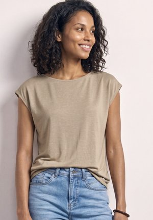 Femme souriante aux cheveux bouclés portant un haut beige à manches courtes rentré dans un jean bleu clair, debout contre un mur pâle.
