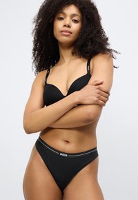 Schwarzer BH mit tief ausgeschnittenen Cups und verstellbaren Trägern, die ein Logo tragen. Passender schwarzer Tanga mit dezentem Taillenlogo und glatter Textur.