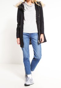 Svart parkas med fuskpäls på huvan, dragkedja och tryckknappar, lager på lager över en grå cowl neck-top och blå jeans. Grå träningsskor.