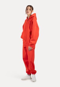 Rode rits hoodie van fleece, gecombineerd met bijpassende joggingbroek. Bevat kangaroo zakken, geribbelde boorden en witte sneakers.