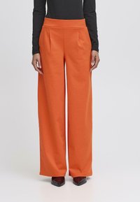 Hoge taille, wijdvallende broek in feloranje stof, met subtiele plooien. Soepele textuur en schone lijnen, gecombineerd met een zwart topje en schoenen.