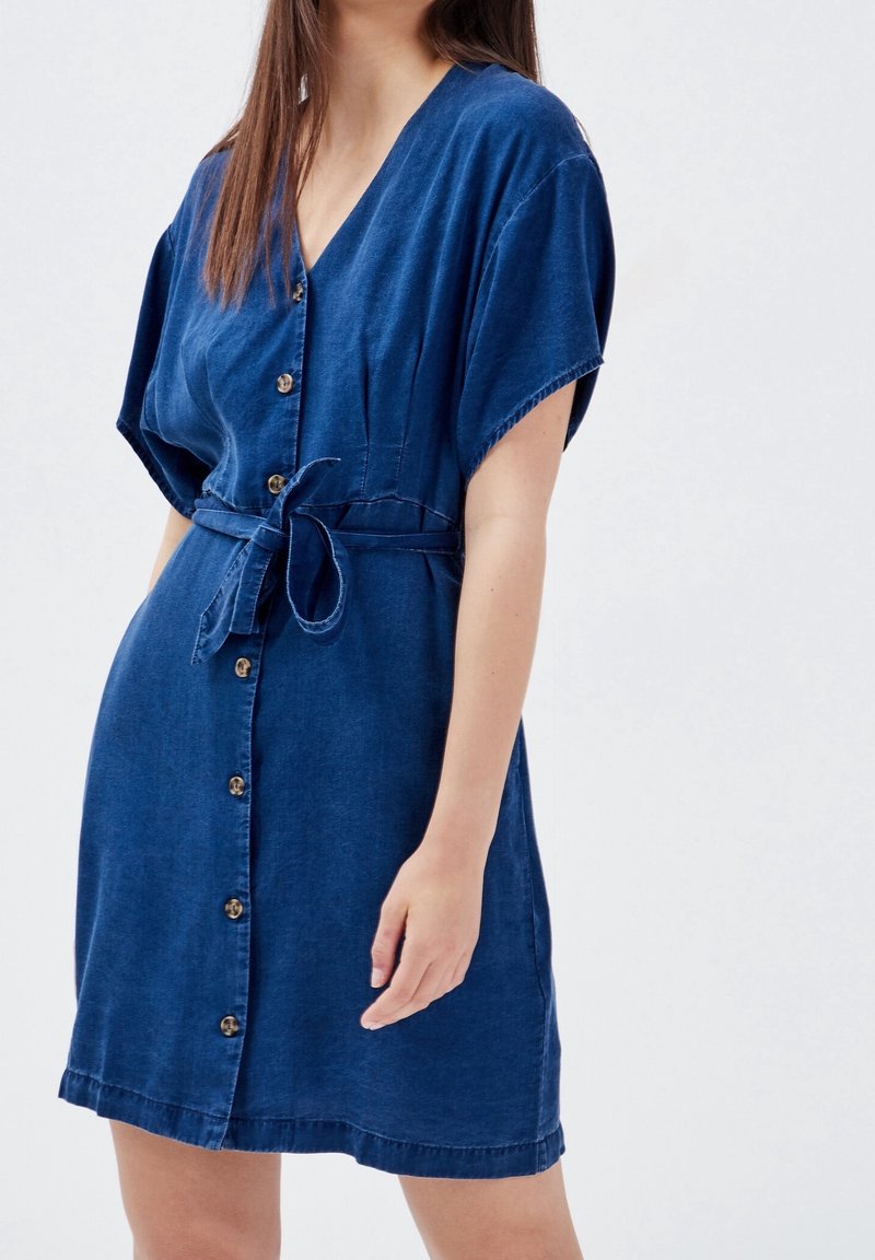 Džínové šaty, tmavě modré, s krátkým rukávem, véčkovým výstřihem, zapínáním na knoflíky vpředu, s úzkým pasem na zavazování, mají volný střih a lem nad koleny.
