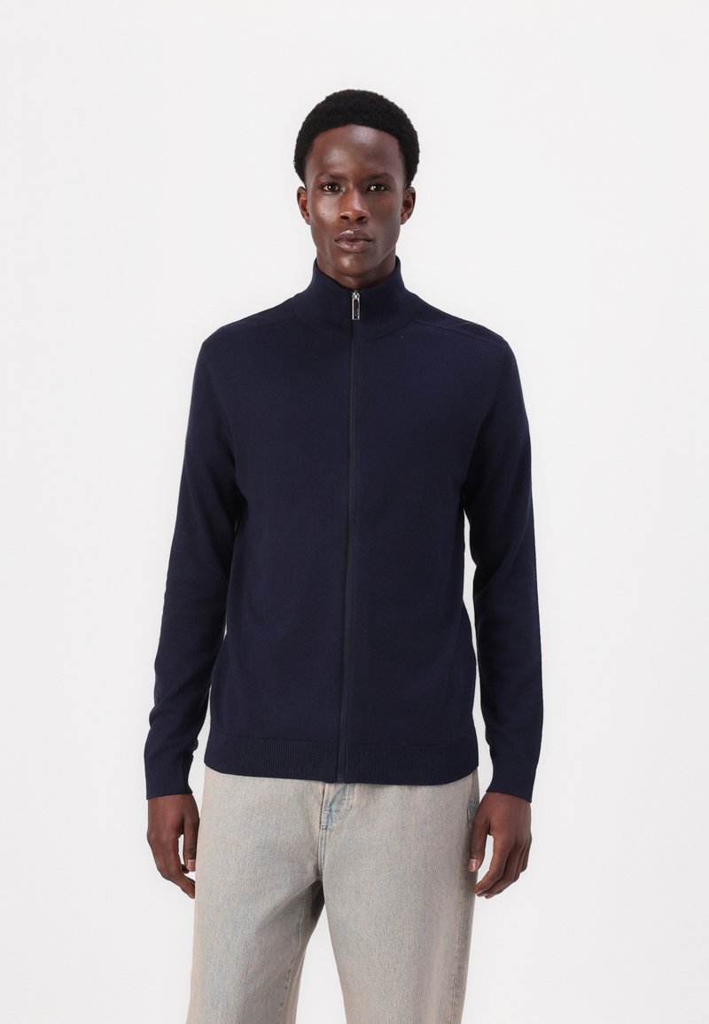 Navy zip-up sweater in a soft knit fabric with a high collar and ribbed hem, paired with light denim pants.