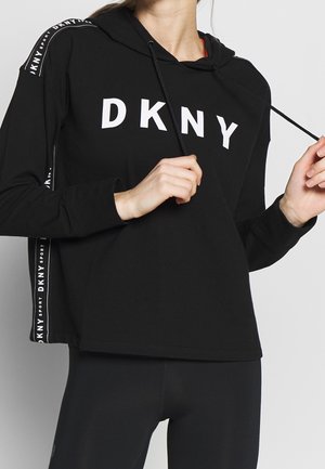 Czarna bluza z kapturem DKNY z białym logo na piersi i taśmą z logo na rękawach, noszona przez osobę regulującą ściągacze.