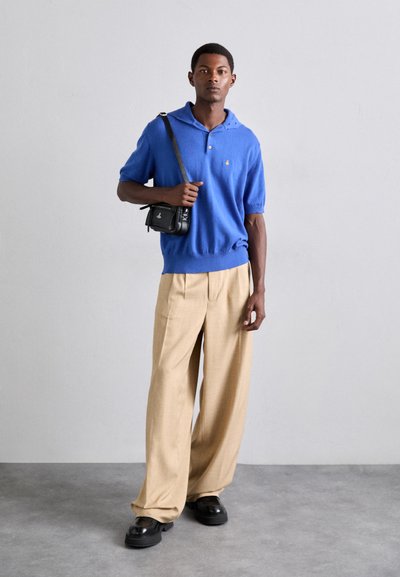 Blå pikétröja med korta ärmar och huva, beige vidbyxor, svart tvärgående axelväska och klumpiga svarta skor. Enfärgad bakgrund.