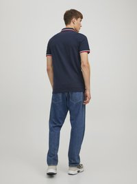 Marineblå polo-shirt med rød og hvid kant ved kraven og ærmerne, parret med løstsiddende blå jeans og grå sneakers.