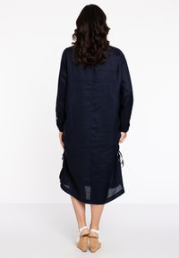 Navyblaues Leinenkleid mit langen Ärmeln, seitlichen Bindebändern und einem leicht geschwungenen Saum. Es hat eine lockere Passform und eine strukturierte Oberfläche.