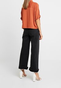 Rusti-oranssi lyhythihainen paita rennolla leikkuulla, yhdistetty korkeavyötäröisiin, mustiin leveälahkeisiin farkkuihin ja valkoisiin paksukorkoisiin sandaaleihin.