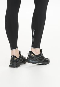 Zwarte legging met contrasterende grijzige accenten en geometrisch patroon aan de zoom, gecombineerd met zwart-grijze sportschoenen met textuuroverlay.