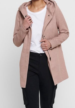 Vrouw die een taupekleurige gewatteerde jas met capuchon sluit over een wit T-shirt, gecombineerd met een zwarte broek, waarbij ze ringen aan haar vingers toont.