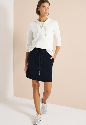 Žena v bílé mikině se šněrováním, černé sukni se stahovacími šňůrkami a stříbrných teniskách, stojící u béžové zdi a dívající se stranou.