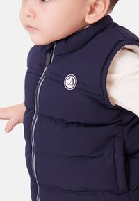 Temno modra puhovka brez rokavov za malčke z zapiranjem na zadrgo in okroglim logotipom. Ima prešite odseke in visok ovratnik za toploto.