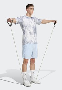 Športové tričko v šedej a bielej farbe s tie-dye vzorom, svetlo modré šortky, čierne odporové gumy, biele topánky a ponožky s čiernymi doplnkami.