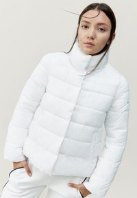 Chaqueta de plumas blanca con un cuello alto, que presenta secciones acolchadas y un bolsillo lateral. Acabado suave, diseño ligero.